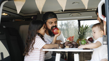 Eine Familie sitzt in einem Van und die Tochter steckt ihrem Vater einen Apfel in den Mund.