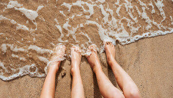 Zwei Menschen mit nackten Füßen sitzen am Strand und halten ihre Füße ins Wasser. Es kommt eine Welle und das Wasser schäumt.
