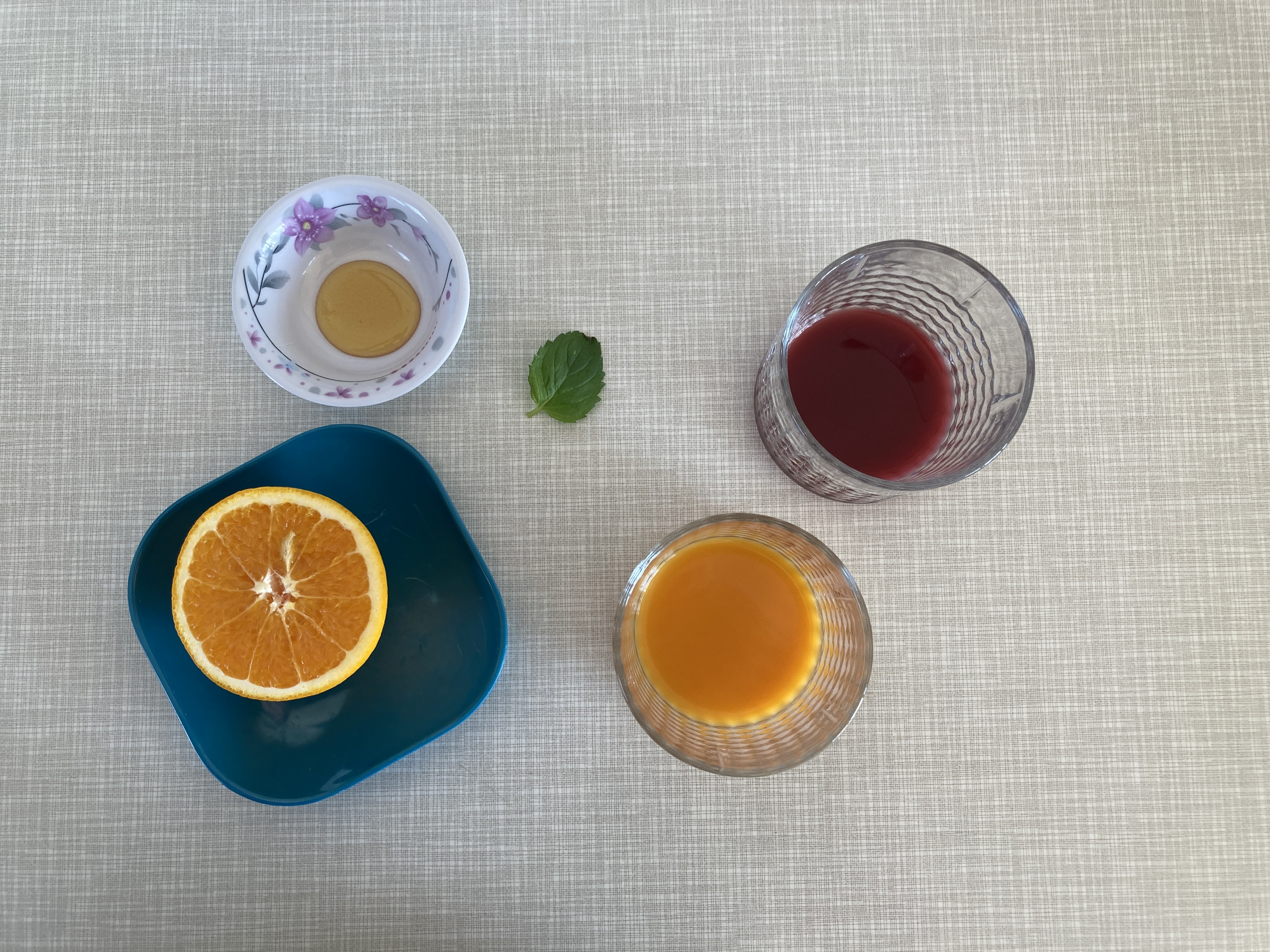 Foto von oben: Auf einem Tisch stehen zwei Gläser mit orangener und roter Limonade. Daneben liegen eine Orange und ein Pfefferminz-Blatt.