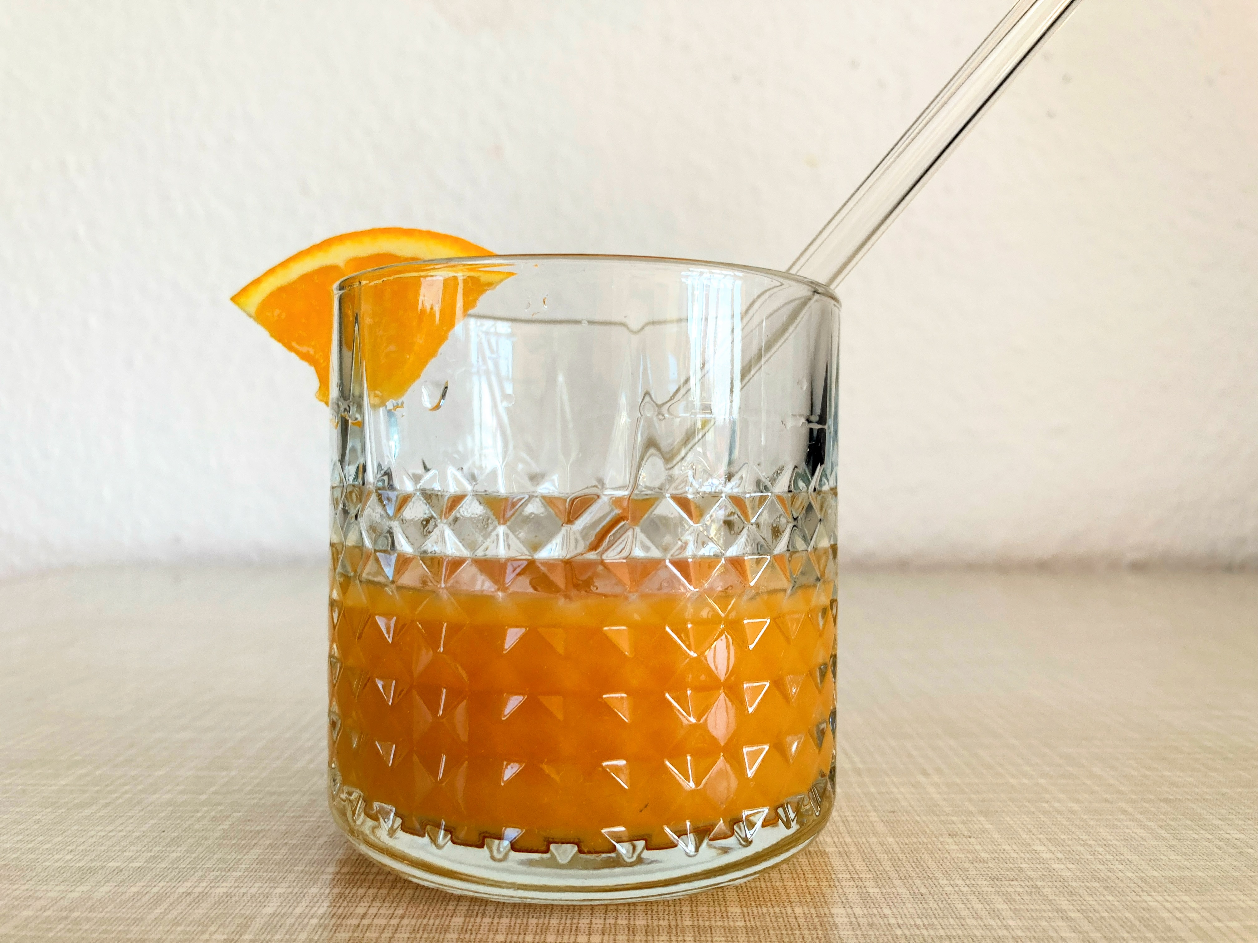 Ein Glas mit einer orangenen Flüssigkeit, einer Orangenscheibe am Rand und einem Strohhalm steht auf einem Tisch.