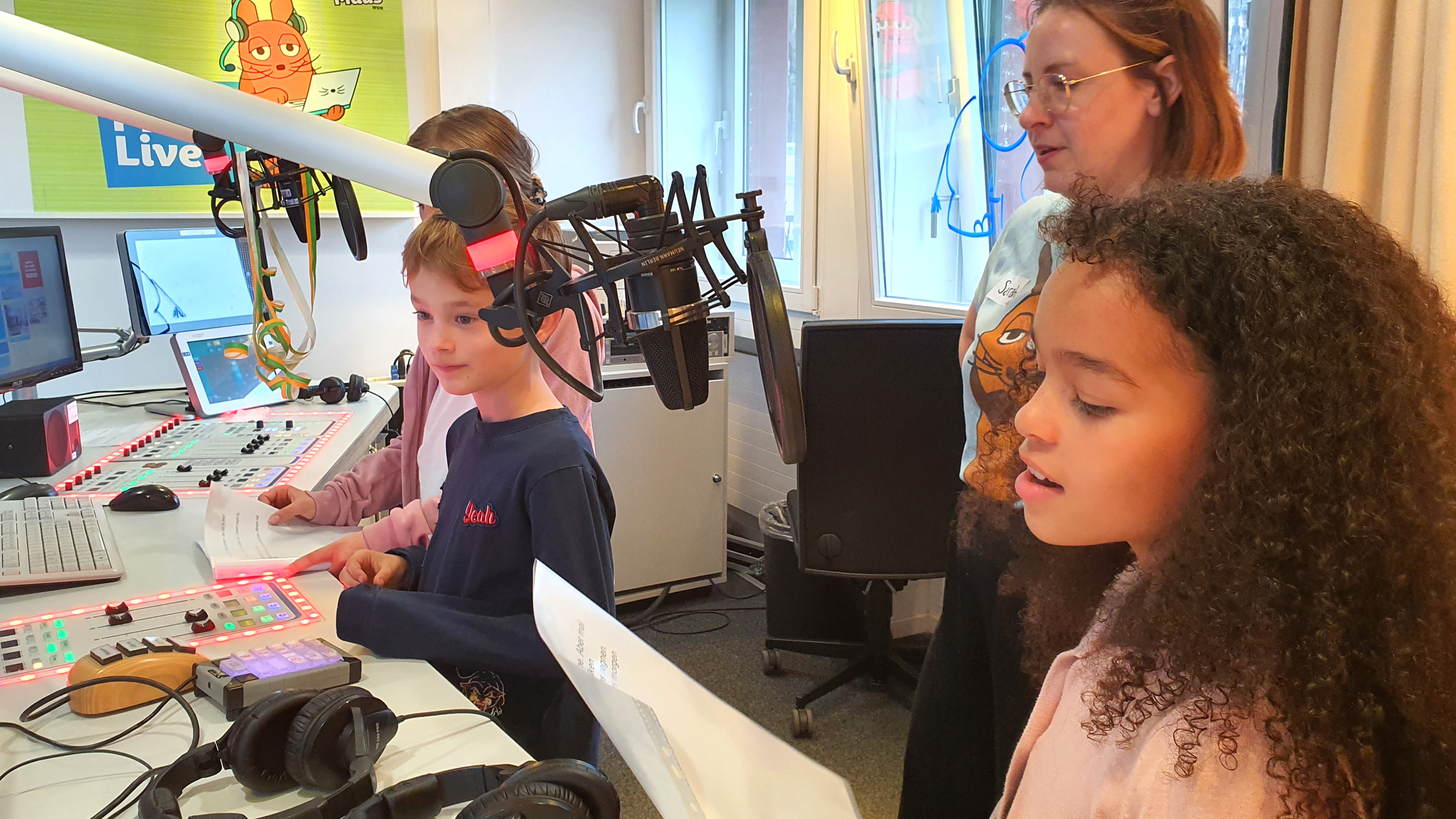 Drei Kinder mit MausLive-Moderatorin Sarah am Sendepult im MausLive-Studio