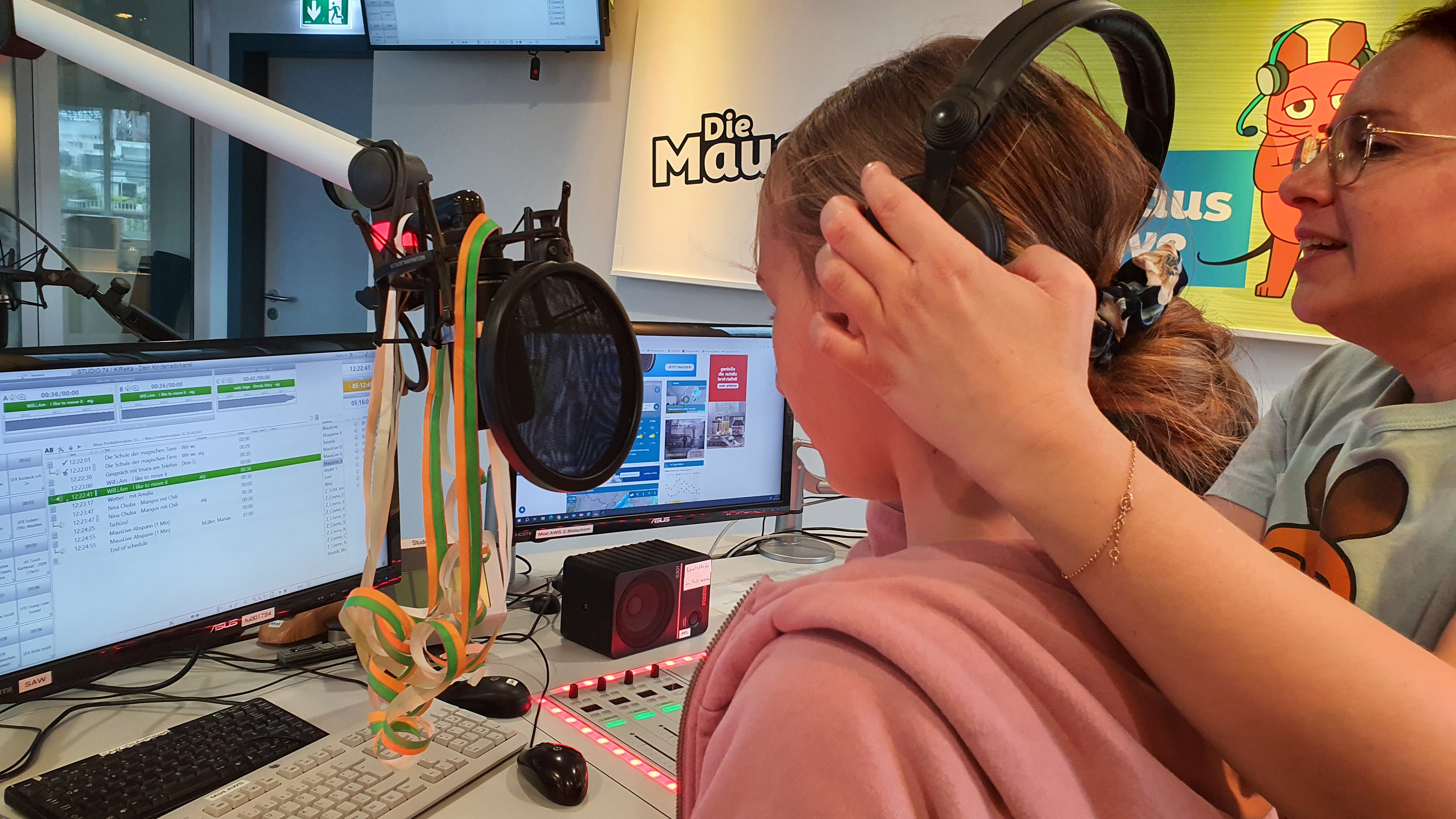 MausLive-Moderatorin Sarah setzt einem Mädchen am Sendepult Kopfhörer auf
