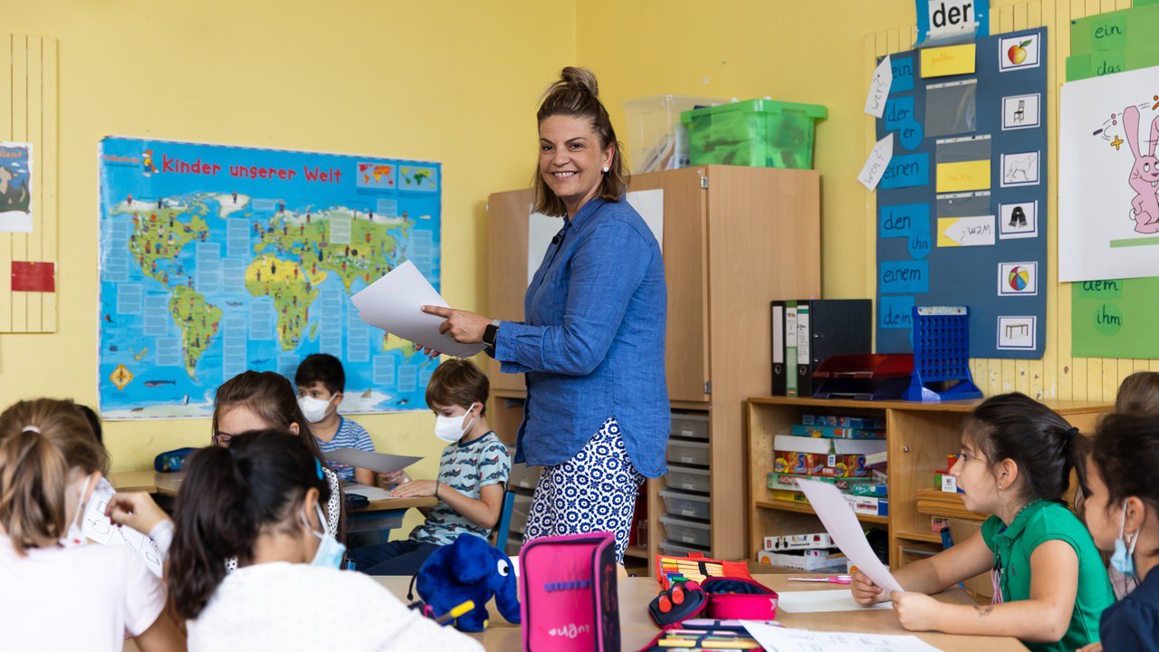 Lehrerin steht lächelnd mit Blättern in der Hand in ihrer Grundschul-Klasse